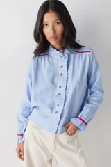 Camisa de lino celeste con cuello alto, volados en la cartera frontal y puños, y detalles de vivos en color rojo en hombros y mangas. Presenta cierre frontal con botones de madera.