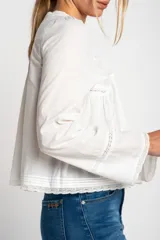 Blusa blanca de algodón con escote en U y mangas largas acampanadas. Presenta detalles de encaje en el escote, el frente y los puños.