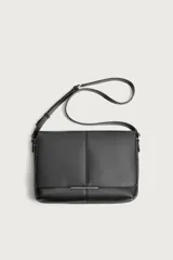 Bandolera de diseño rectangular en color negro con acabado texturizado. Cuenta con una solapa frontal con detalle metálico, cierre imantado y asa de hombro ajustable. El interior incluye un bolsillo plano y otro con cierre, además de un bolsillo plano adicional en la parte frontal.