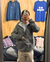 Rompeviento gris de estilo canguro con capucha, cuello alto y bolsillo frontal tipo sobre con cierre. Presenta puños elásticos y diseño funcional impermeable.