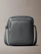 Bolso bandolera Calvin Klein color gris oscuro, de cuero sintético con textura granulada. Tiene correa ajustable, cierre superior con cremallera, bolsillo frontal con cremallera y logo de la marca grabado en la parte inferior.