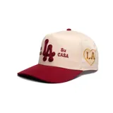 Gorra GQH de visera plana, color blanco con visera bordó. Presenta bordados frontales en relieve con las letras "LA" y el texto "Mi Casa Su Casa" en color bordó.