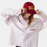 Camisa de manga larga con cuello y botones frontales, corte boxy, en color blanco con estampado de cuadrícula fina en tono rosado. Presenta un pequeño logo bordado en rojo en el pecho.