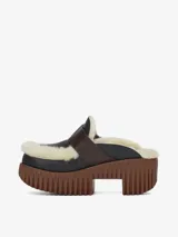 Zapato tipo mule con plataforma track de color marrón y capellada de cuero negro. Cuenta con forro de lana de oveja (borreguito) visible en los bordes e interior, y una correa de cuero marrón con hebilla metálica sobre el empeine.