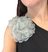 Broche en forma de flor grande confeccionado en organza traslúcida con acabado brillante. Cuenta con un sistema de sujeción versátil que permite usarlo tanto en prendas de vestir como en el cabello.