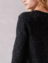 Cardigan de punto con textura tipo tweed en color negro con destellos dorados. Presenta escote en V, cierre frontal con botones metálicos decorativos y bolsillos frontales con solapa. Posee terminaciones acanaladas en cuello, puños y ruedo.