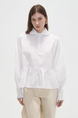 Camisa blanca de poliéster, con cuello redondo abotinado con solapa fruncida, manga larga con puños elásticos y corte con volados en la parte inferior.