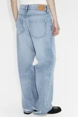Jean de corte wide leg con diseño de cinco bolsillos, confeccionado en denim de algodón rígido. Presenta un lavado claro con efecto desgastado y un detalle de rotura en la rodilla izquierda. Cuenta con cintura clásica, cierre de botón y bragueta.