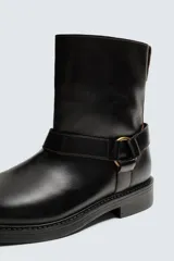 Bota de caña media fabricada en piel color negro, con detalle de arnés y hebilla metálica lateral. Presenta horma cuadrada y suela a tono con ligero tacón.