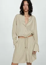 Camisa color beige de corte crop, confeccionada en mezcla de lino y lyocell. Tiene cuello camisero, mangas largas con puños abotonados, lazo ajustable en la cintura y cierre frontal con botones.