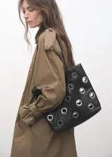 Bolso tipo tote de color negro, confeccionado en poliuretano, con diseño de ojales metálicos plateados distribuidos por toda la superficie y asas largas para llevar al hombro.
