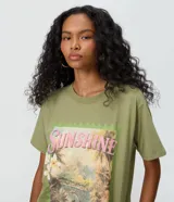 Remera verde militar de algodón, de corte largo y diseño asimétrico. Presenta mangas cortas, cuello redondo y una estampa frontal con un paisaje tropical, flores y las inscripciones "Sunshine" y "Having a Good Time".