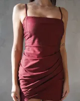 Vestido corto de fiesta color bordó con acabado brillante, escote recto y breteles finos. Presenta un diseño ajustado con detalle de drapeado cruzado en la falda.