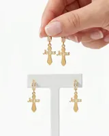 Caravanas de acero dorado con diseño de cruz que incorpora la palabra 'Jesus' en el centro. Cuentan con un aro tipo argolla decorado con pequeñas piedras brillantes.