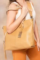 Bolso tipo tote de cuero color beige con textura croco en tono camel. Tiene doble asa corta y correa larga desmontable con diseño de rombos. Incluye flecos decorativos, herrajes metálicos y cierre superior.