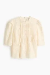 Top de encaje color crema con ribetes festoneados, cuello de volados, mangas cortas abullonadas, alforzas en el frente y cierre oculto en la parte posterior.