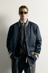 Cazadora bomber estilo universitario, de corte relaxed fit y tejido técnico azul marino, con cuello, puños y dobladillo de rib elástico a rayas amarillas. Cuenta con bolsillos delanteros de plastrón y cierre frontal con botones a presión.