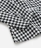 Blazer de sastrería confeccionado en tejido tweed con estampado pied de poule en blanco y negro. Presenta cuello con solapa, cierre frontal con un botón, mangas largas y bolsillos frontales con solapa.