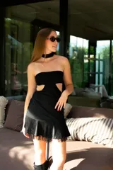 Vestido corto negro con diseño cut-out en la cintura, estilo strapless y ruedo decorado con flecos largos.
