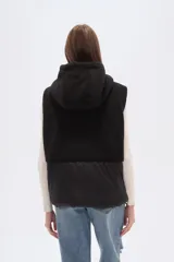 Chaleco con capucha, diseño bicolor que combina textura de corderito en la parte superior y material técnico liso en la parte inferior. Cuenta con cierre frontal y botones a presión.