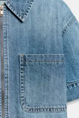 Camisa de denim de corte recto y manga corta, con cuello solapa, cierre frontal con cremallera y dos bolsillos delanteros de plastrón.
