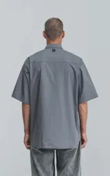 Camisa de manga corta de poplin, color gris, con silueta oversize. Presenta dos bolsillos delanteros, uno con solapa y otro con ojal y botón.