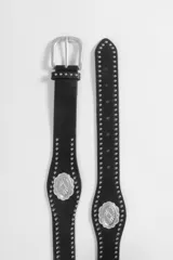 Cinturón de cuero vegano negro con diseño de estilo western, decorado con tachas metálicas plateadas a lo largo de toda la pieza y apliques de medallas ovaladas grabadas.