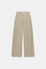 Pantalón de algodón color beige con tiro medio, cintura elástica ajustable con cordón y perneras anchas con pliegues delanteros y bolsillos laterales.
