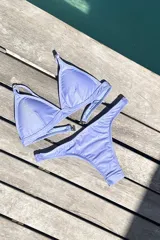 Conjunto de bikini color lila con acabado brillante. El top es de estilo triangular con breteles regulables y cierre de gancho en la espalda. La bombacha es de corte vedetina con laterales fruncidos.