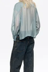 Camisa de corte boxy en color celeste, confeccionada en tejido semitransparente con apliques de pequeñas piedras brillantes. Posee cuello camisero, cierre frontal con botones y mangas largas con puños con sardineta.