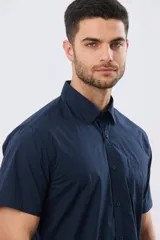 Camisa lisa de algodón azul marino, de manga corta, con cuello camisero, cierre frontal de botones y un bolsillo aplicado en el pecho.