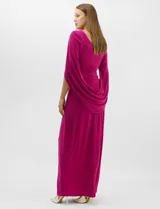 Vestido largo de seda fría color fucsia, con escote cruzado drapeado y apliques de pedrería en los hombros. Presenta mangas largas abiertas estilo capa y un drapeado lateral en la cintura que estiliza la silueta.