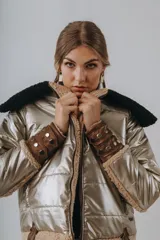 Campera puffer metalizada en tono dorado, con interior y detalles en corderito beige. Presenta capucha negra de corderito, cierre frontal y puños ajustables con tiras de cuero y tachas metálicas.