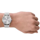 Reloj de pulsera analógico con caja de acero inoxidable y malla de eslabones plateada. Presenta una esfera minimalista en tono plateado con acabado sunray, índices de bastón, números romanos en posiciones clave, ventana de fecha y el logo de la marca en la parte superior.