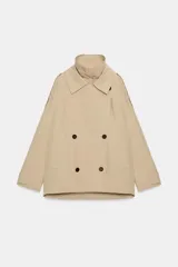Trench corto oversize color beige con cuello subido de efecto doble, cierre cruzado con botones oscuros y bolsillos de vivo delanteros.