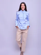 Camisa de algodón color celeste, con cuello alto, cierre frontal de botones y detalles de volados verticales a lo largo de la botonera. Presenta mangas largas con puños ajustados y terminación con volados.