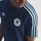 Remera de algodón azul marino con cuello redondo y mangas cortas. Presenta el logo de Adidas Trefoil bordado en blanco en el lado derecho del pecho y el escudo de la selección alemana de fútbol en el lado izquierdo. Incluye las icónicas tres tiras de la marca en color celeste claro a lo largo de los hombros y mangas.