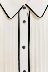 Vestido midi blanco, sin mangas, con cuello camisero y cierre frontal de botones. Presenta un plisado vertical en todo el cuerpo y ribetes negros a contraste en el cuello, tapeta de botones y borde de la abertura inferior. Se complementa con un cinturón negro de hebilla dorada.