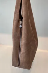 Bolso tipo tote de gamuza en color verde oliva, con diseño minimalista y asa integrada. Presenta una placa metálica con el logo de la marca en la parte inferior central y detalles de textura en el asa.