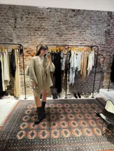 Ruana tejida a mano en lana merino color beige, con textura de punto y detalle de flecos en el ruedo. Presenta sutiles destellos brillantes integrados en el tejido.