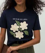 Remera de algodón color blanco, con cuello redondo y manga corta. Presenta un estampado con la frase 'Soy flor. Soy latina' y flores bordadas en relieve con textura tejida.