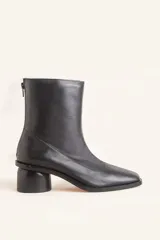 Bota de caña media confeccionada en cuero vacuno negro, con diseño minimalista y cierre trasero. Presenta un taco cilíndrico de madera tallada y punta cuadrada.