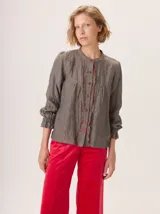 Camisa de algodón en tono gris azulado, con cuello redondo, cierre frontal de botones y detalles de volados con costuras en contraste color rojo. Presenta mangas largas con puños elásticos y diseño fluido.
