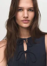 Vestido corto sin mangas de efecto denim en color azul marino, con cuello redondo y tirantes anchos. Presenta detalles de lazos en la parte delantera y diseño de tablas en la falda.