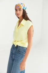 Camisa de lino color amarillo manteca, sin mangas, con cuello clásico y cierre frontal con botones. Presenta un diseño corto con nudo en el ruedo.