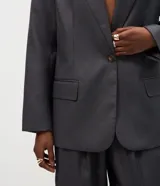 Blazer de sastrería color gris oscuro, con cuello solapa, cierre frontal de un botón y bolsillos laterales con tapa. Presenta un diseño estructurado con mangas largas y forro interno.