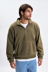 Buzo de polar verde militar con medio cierre frontal, cuello alto y bolsillo tipo canguro. Presenta un pequeño logo bordado en el pecho.