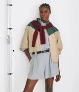 Campera rompeviento de diseño tricolor con bloques de color en verde, beige y blanco. Presenta cuello alto, cierre frontal con cremallera, mangas raglán y puños ajustados. El ruedo incluye un cordón regulable para un ajuste personalizado.