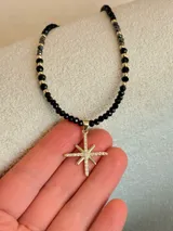 Collar corto de cuentas negras facetadas con cuentas metálicas doradas intercaladas, con un dije colgante en forma de estrella de ocho puntas dorada con pavé de circones blancos.