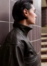 Chaqueta bomber de cuero ovino color marrón oscuro, con cierre frontal de botones, bolsillos con solapa en el pecho y cuello alto con broches.